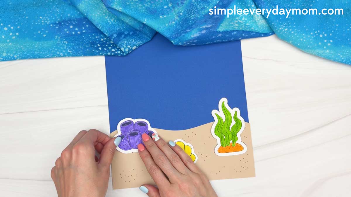 Hands attaching a purple coral decoration to the sandy ocean background for the whale shark craft scene.