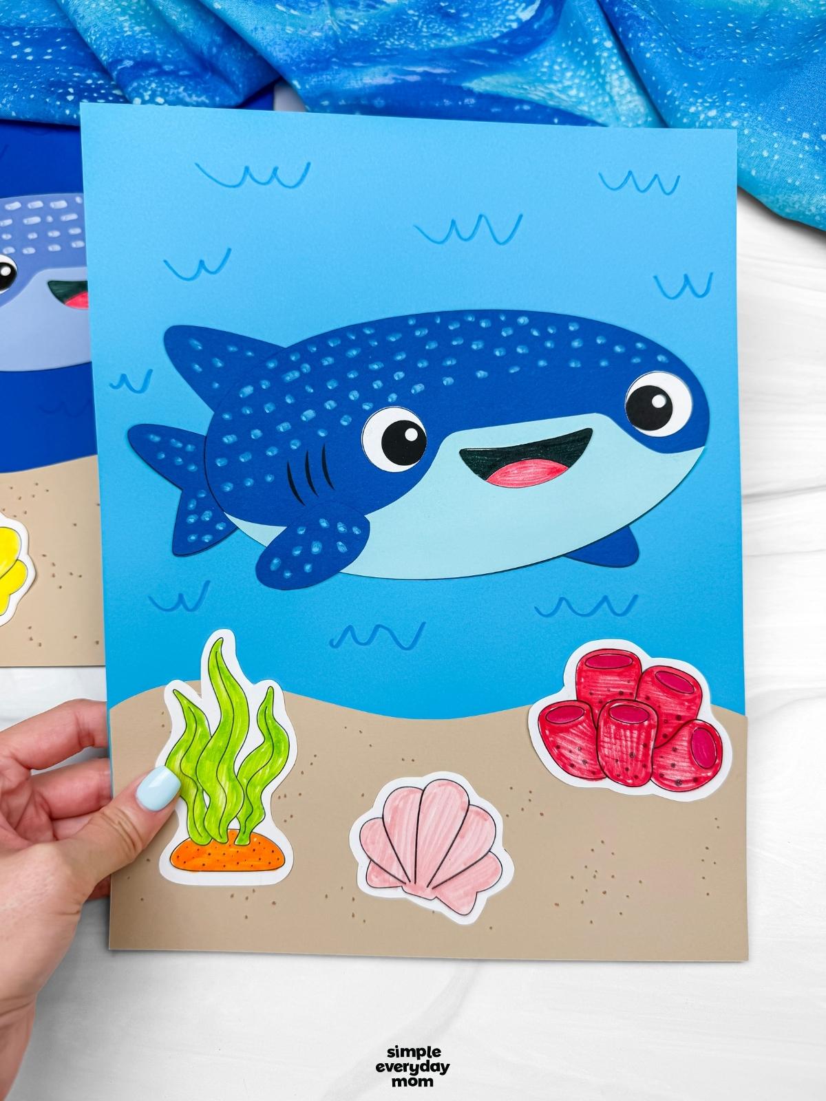 A completed Craft showing a whale shark paper craft on an ocean themed background with seaweed, coral, and shell decorations.