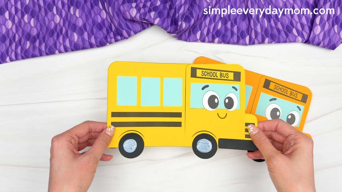 Hands holding the completed bus shape craft made from simple geometric shapes with a smiling school bus face.
