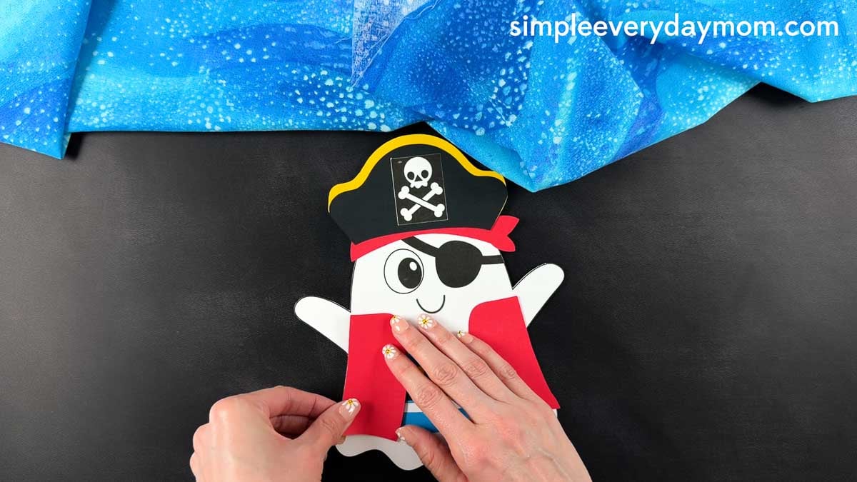 Hands gluing the pirate coat pieces to each side of the ghost's body below the arms.