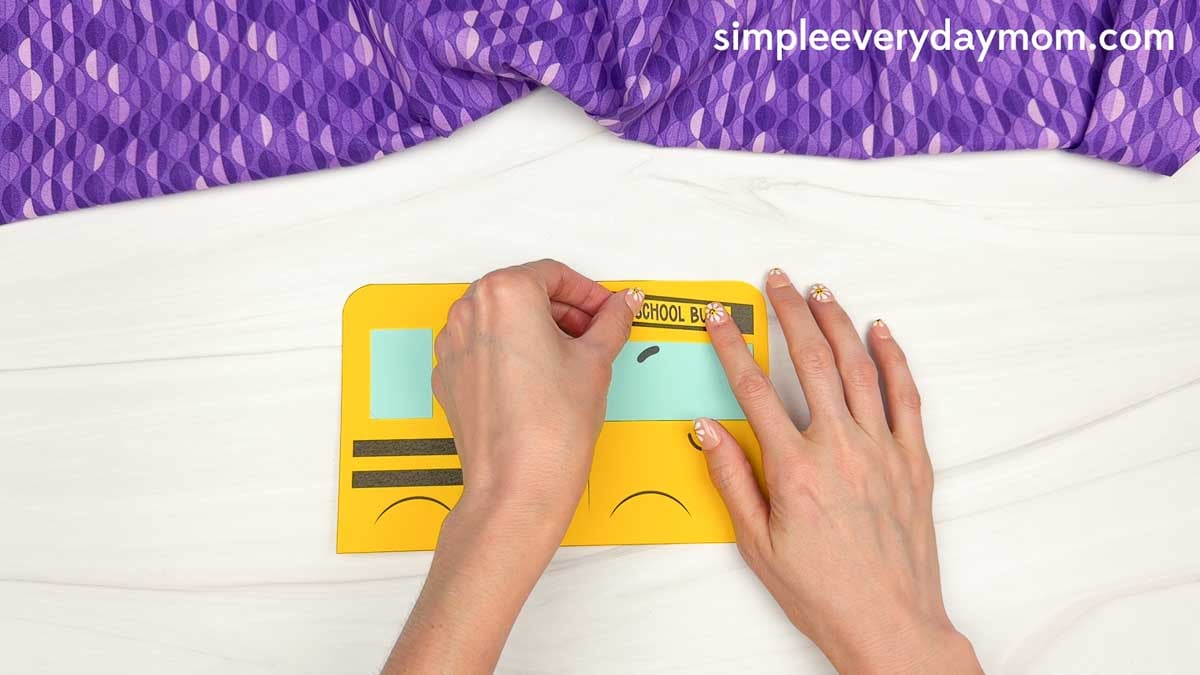 Hands placing the school bus sign piece above the front window on the bus shape craft template.