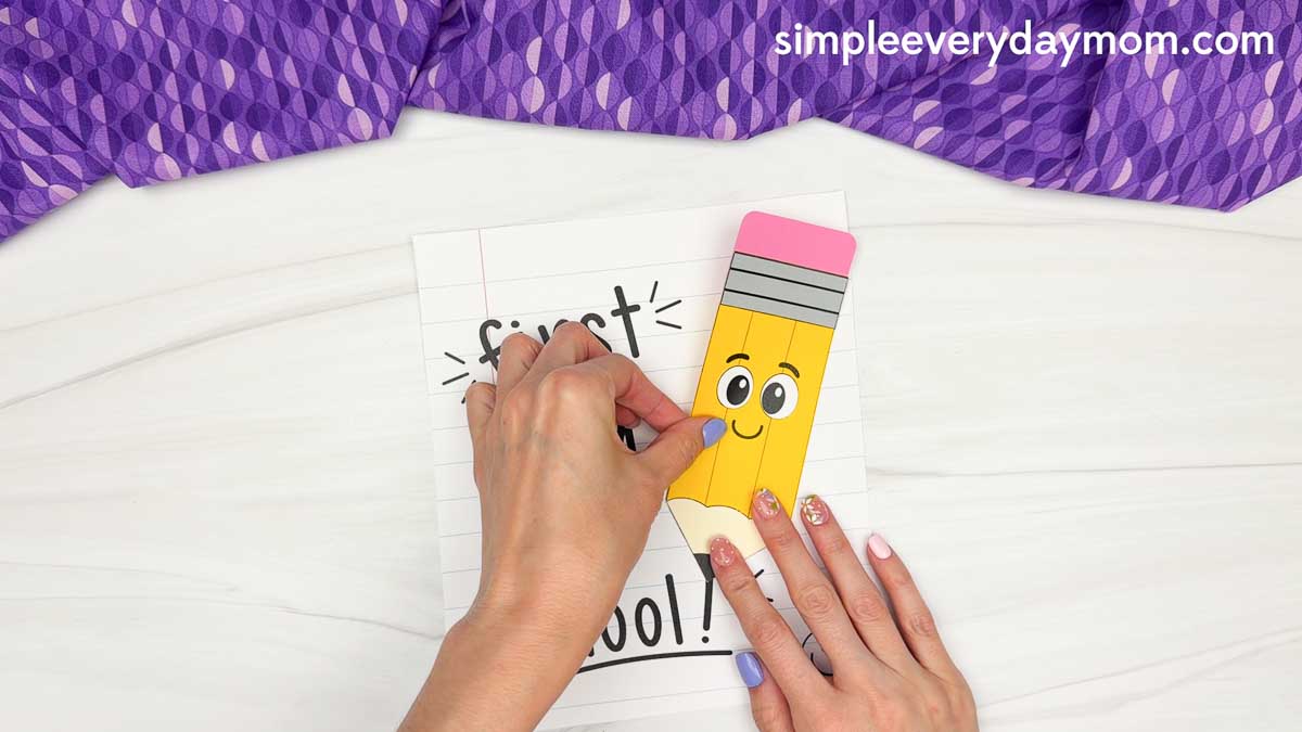 Hands attach the smiling pencil craft to the front of the first day of school notebook paper.