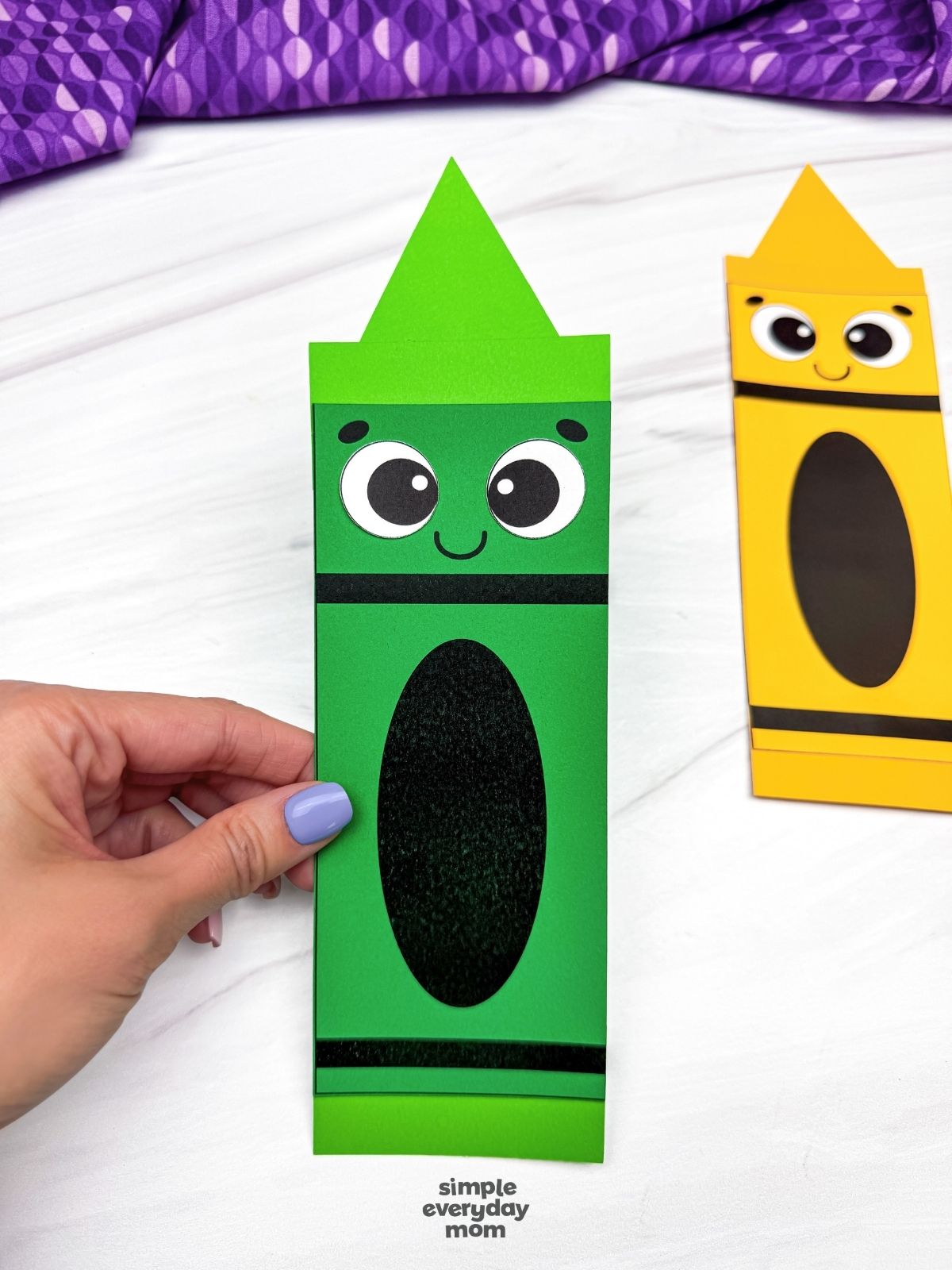 Hands holding the completed green crayon shape craft with a yellow crayon shape craft in the background.