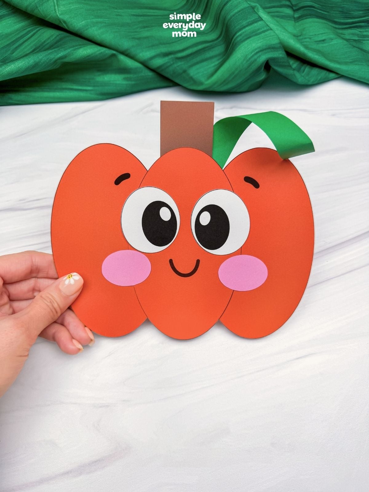 Hands holding a completed Pumpkin Shape Craft with a smiling face, stem, and curled vine.