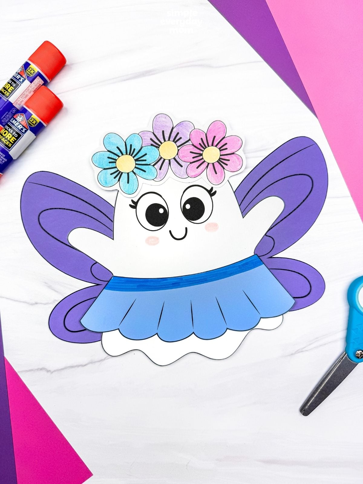A completed craft of the Fairy Ghost Disguise Craft with purple fairy wings, a flower crown, and a blue skirt surrounded by craft supplies.