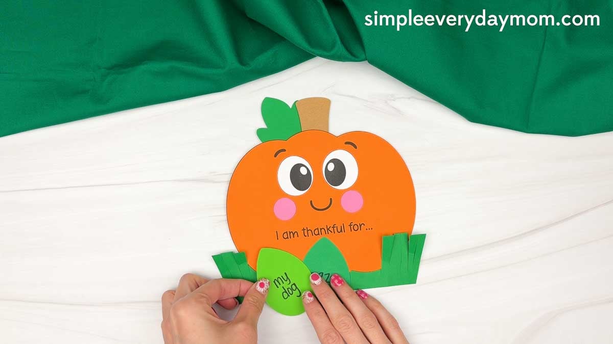 Hands sliding the thankful leaf pieces into the grass pockets below the thankful pumpkin craft.
