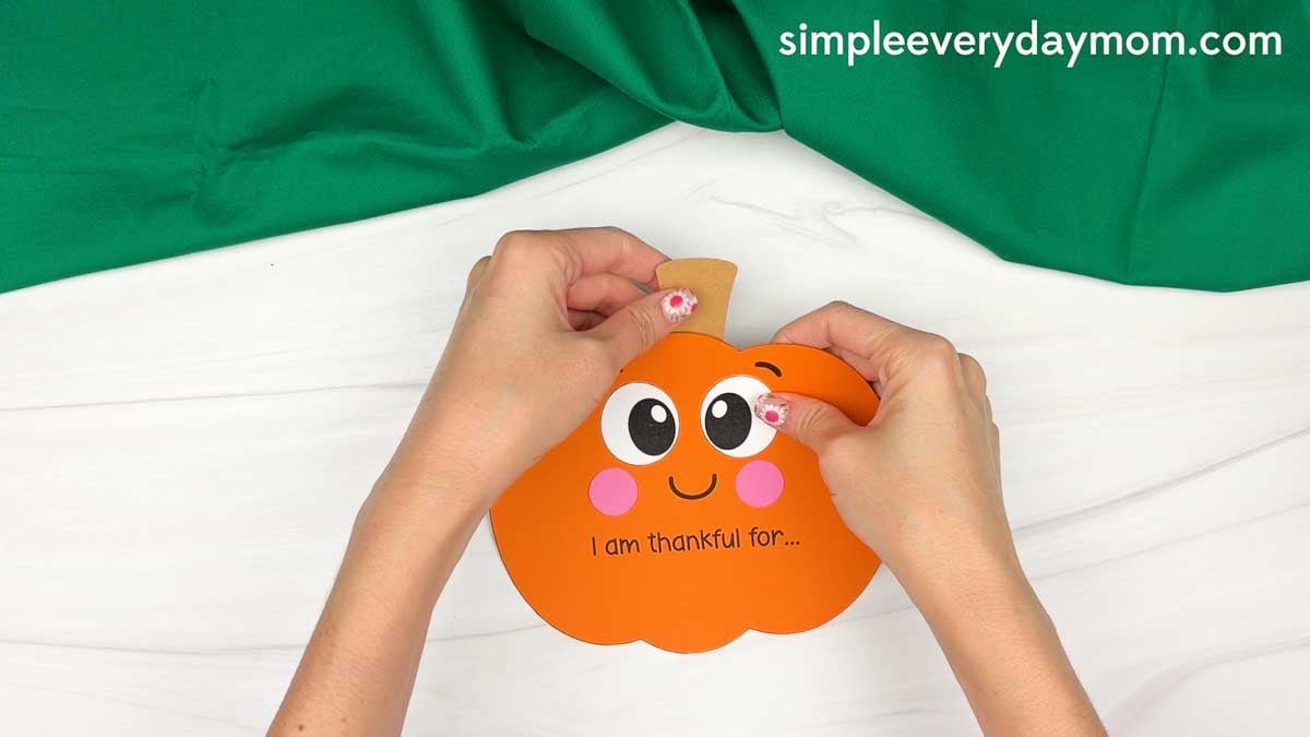 Hands gluing the stem to the top center of the thankful pumpkin above the face.