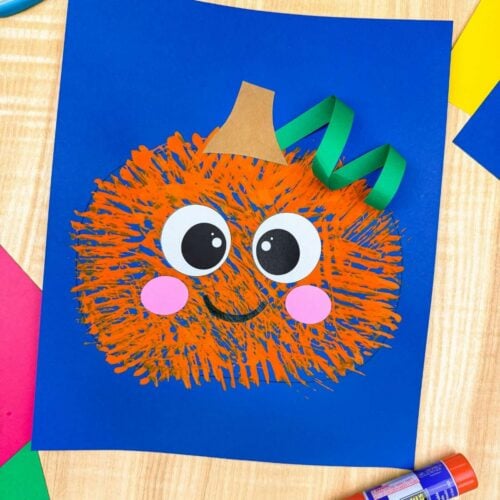 A completed craft of the Fork Painted Pumpkin Craft displayed on blue paper with colorful craft supplies around it.