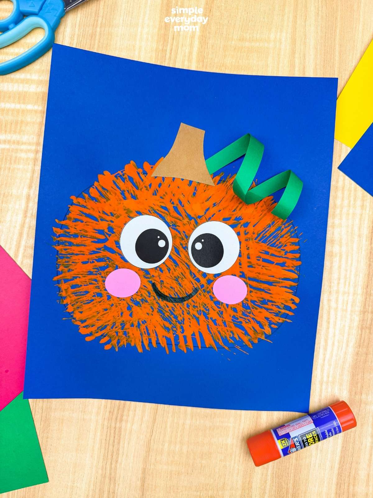 A completed craft of the Fork Painted Pumpkin Craft displayed on blue paper with colorful craft supplies around it.