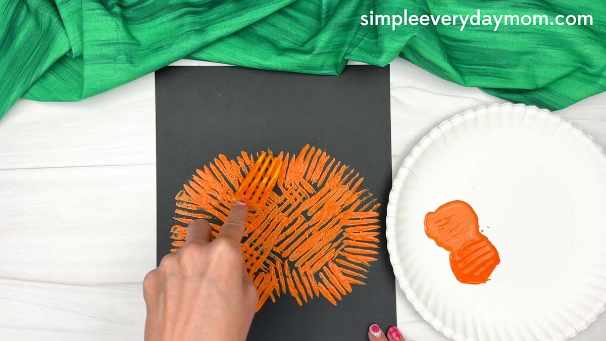 Hands using a fork to paint the pumpkin shape onto black paper with orange paint on a paper plate nearby.