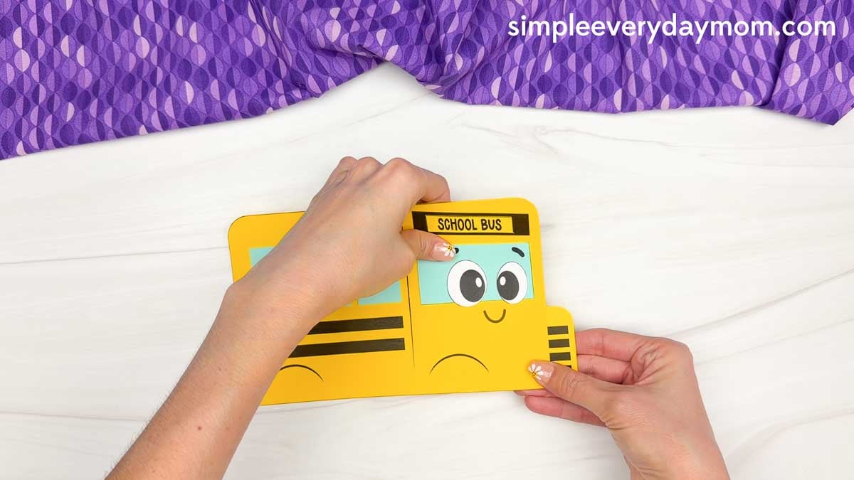 Hands gluing the face piece below the school bus sign on the bus shape craft template.