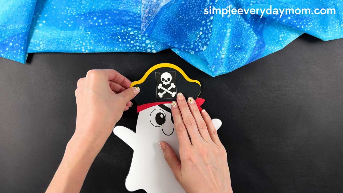 Hands gluing the completed pirate hat to the top of the ghost's head above the pirate bandana.