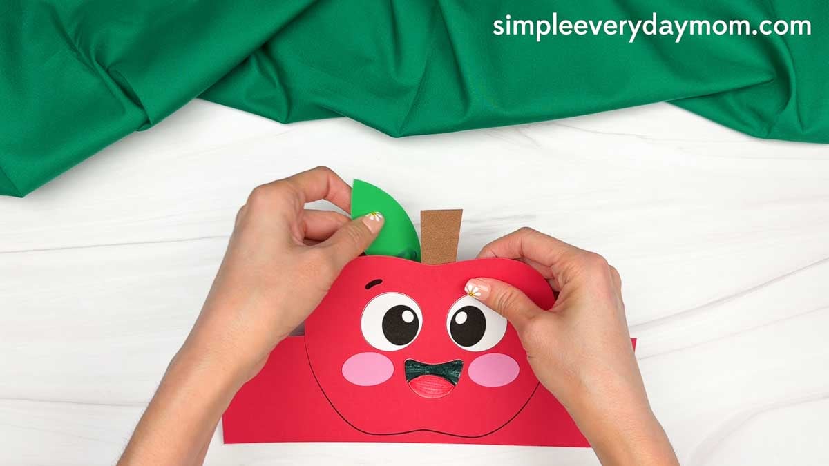 Hands gluing the leaf piece behind the top of the apple headband template beside the stem.