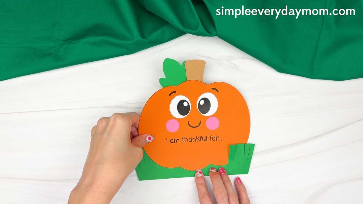 Hands gluing the thankful pumpkin onto the paper grass backing at the bottom edge.