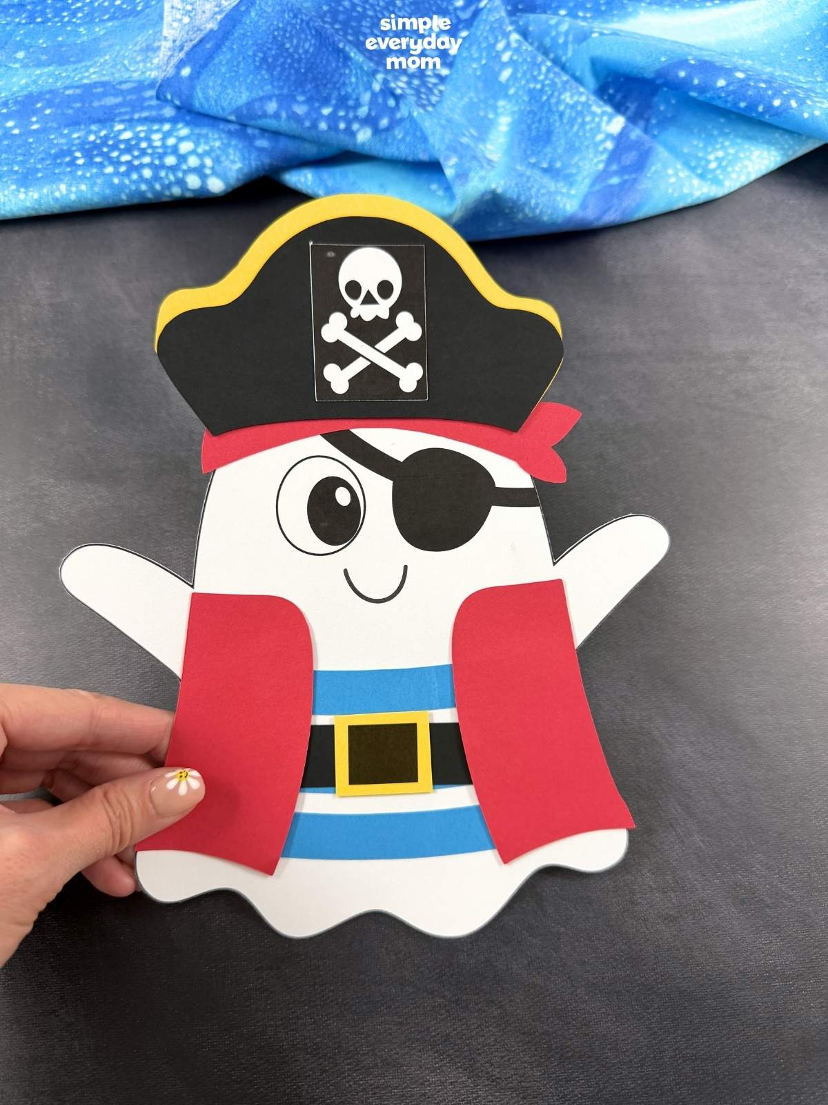 Hands holding the completed pirate ghost disguise craft with a pirate hat, eye patch, striped shirt, belt, and red pirate coat.