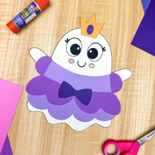 A completed craft of the Princess Ghost Disguise Craft featuring a layered princess dress, bow, sleeves, and crown displayed with colorful paper, scissors, and a glue stick.