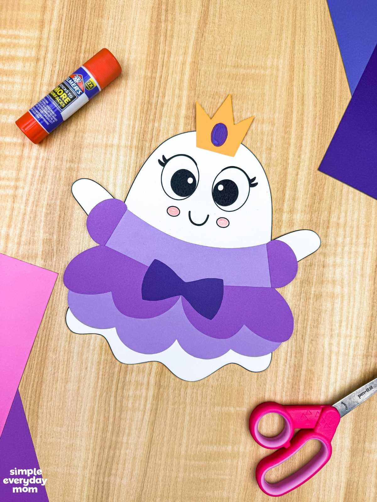 A completed craft of the Princess Ghost Disguise Craft featuring a layered princess dress, bow, sleeves, and crown displayed with colorful paper, scissors, and a glue stick.