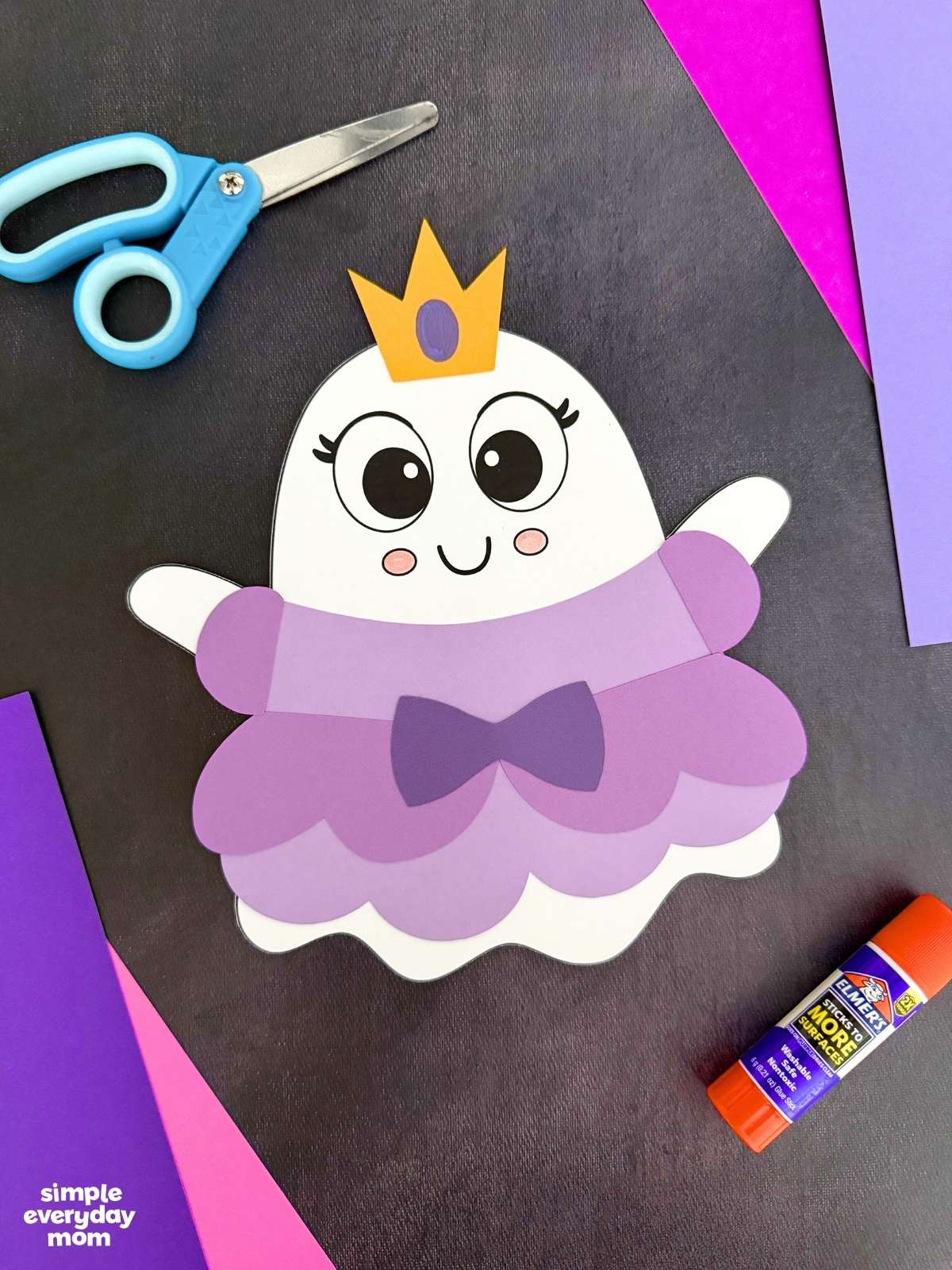 A completed craft of the Princess Ghost Disguise Craft featuring a layered princess dress, bow, sleeves, and crown displayed with colorful paper, scissors, and a glue stick.