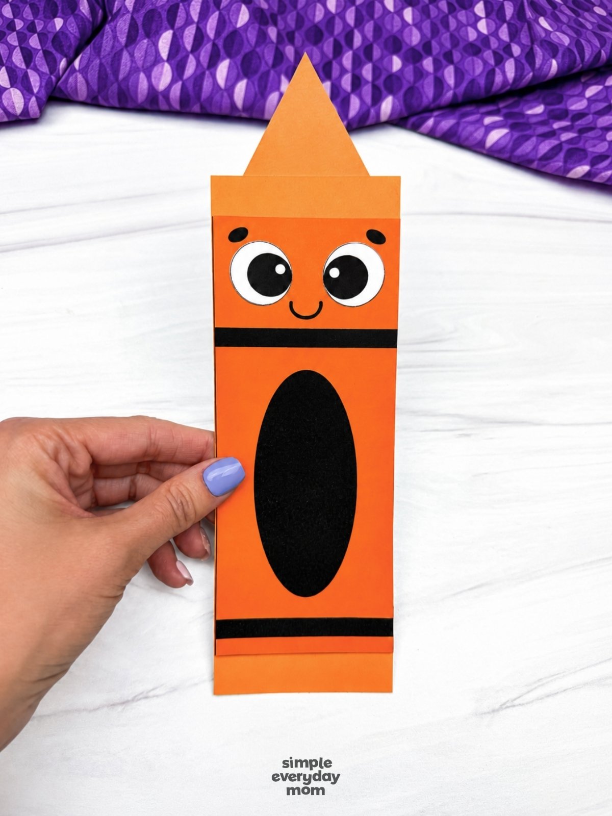 Hands holding the completed orange crayon shape craft with a smiling face.