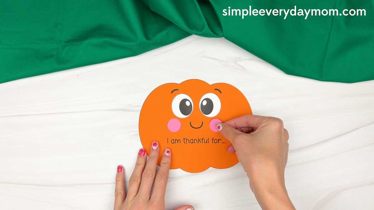Hands gluing the left cheek onto the thankful pumpkin face below the eye to complete the cheeks.