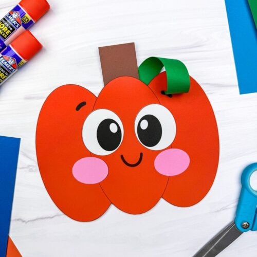 A completed Craft of a smiling Pumpkin Shape Craft displayed with scissors, glue sticks, and colorful paper.