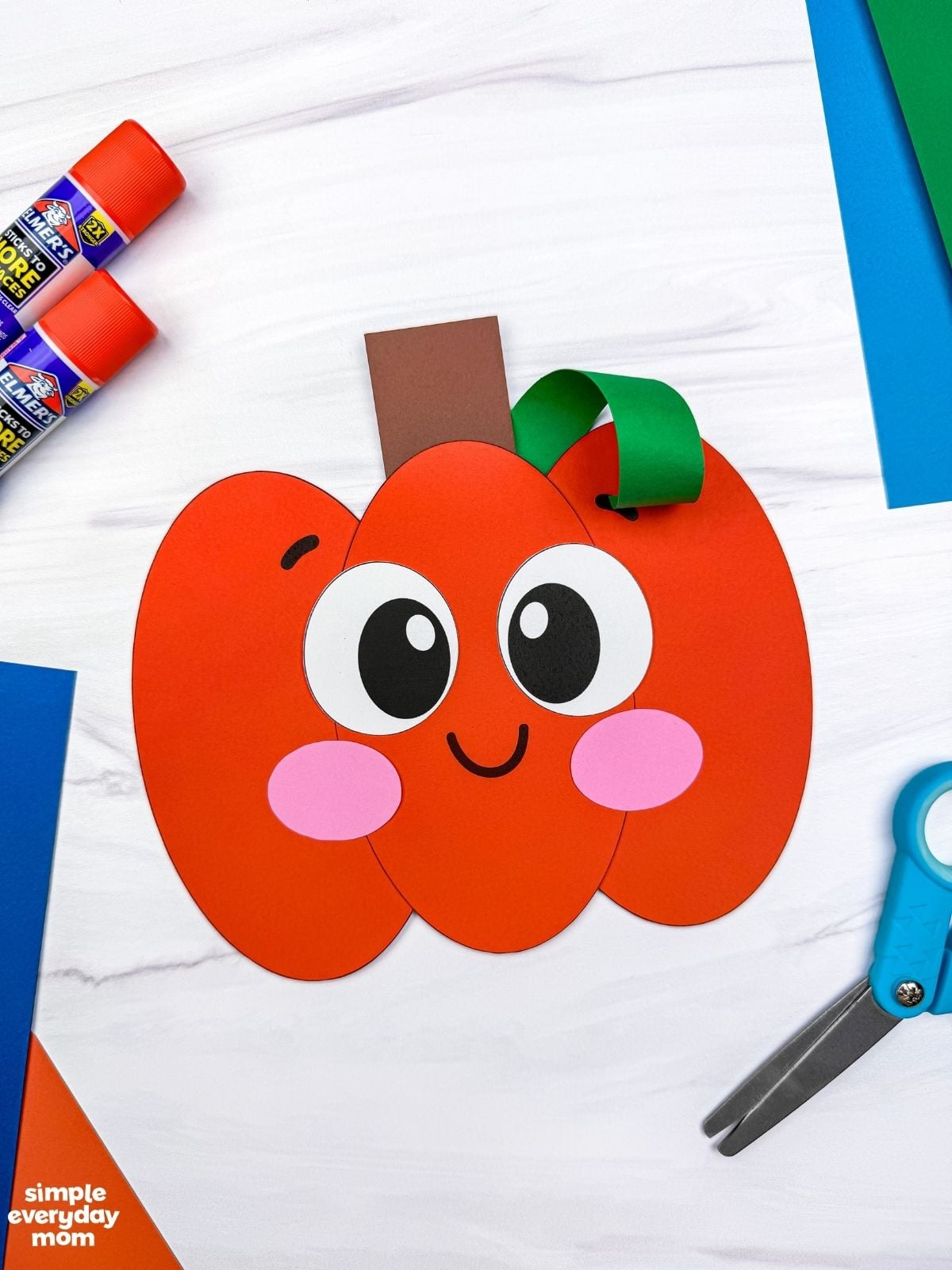 A completed Craft of a smiling Pumpkin Shape Craft displayed with scissors, glue sticks, and colorful paper.