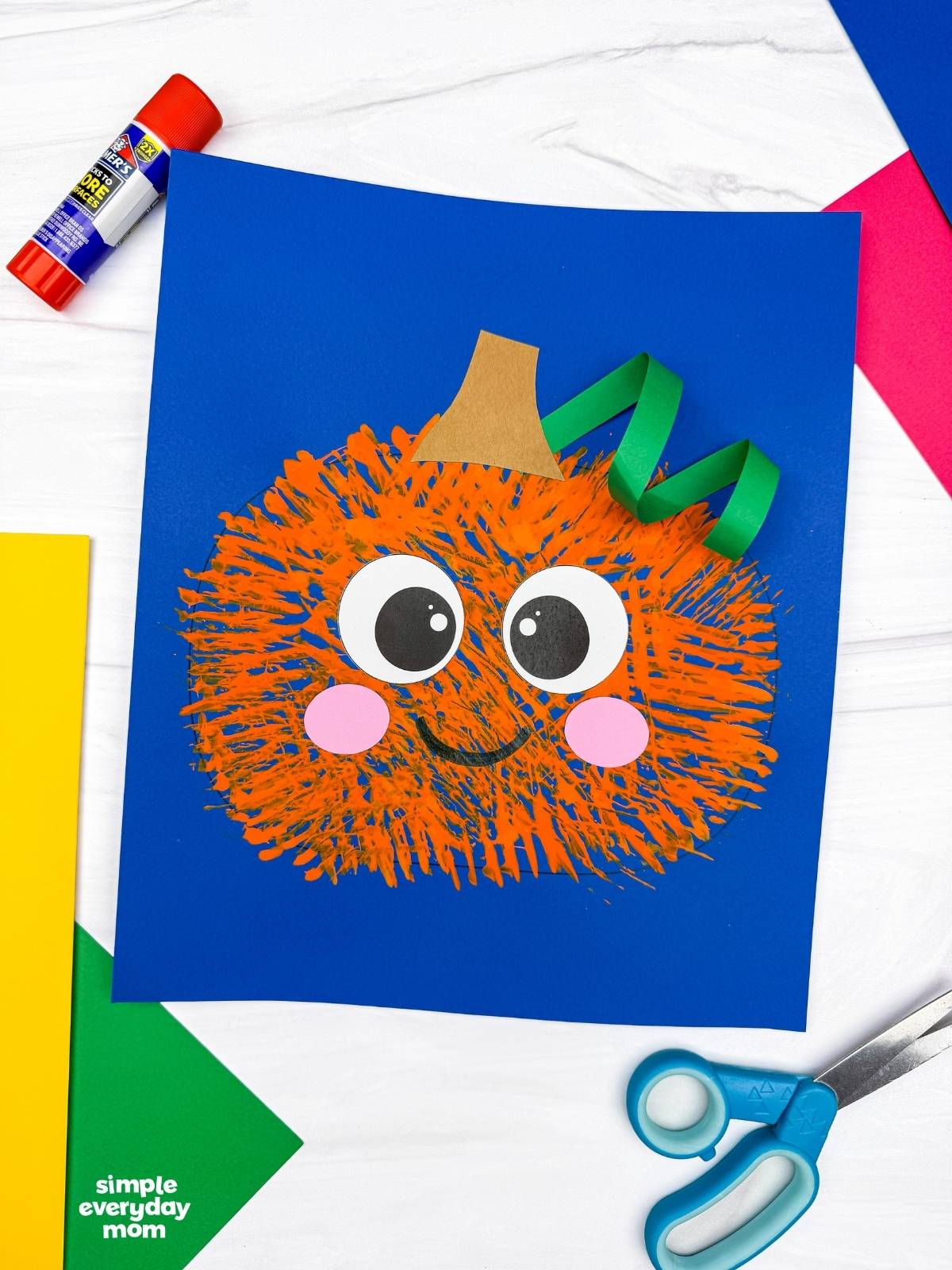 A completed craft of the Fork Painted Pumpkin Craft displayed on blue paper with craft supplies around it.
