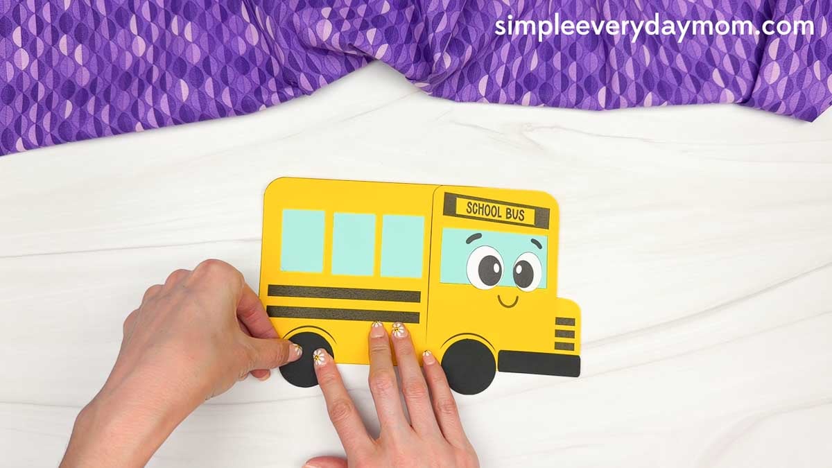 Hands attaching the wheel pieces to the bottom of the bus shape craft template.