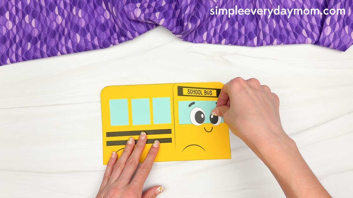Hands placing the right eye onto the bus face below the school bus sign on the bus shape craft template.