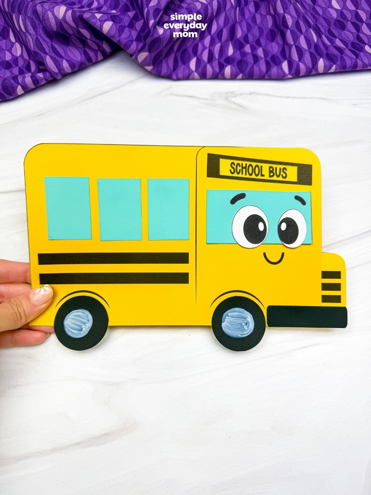 Hands holding the completed bus shape craft with a smiling school bus face and colorful geometric shape windows.