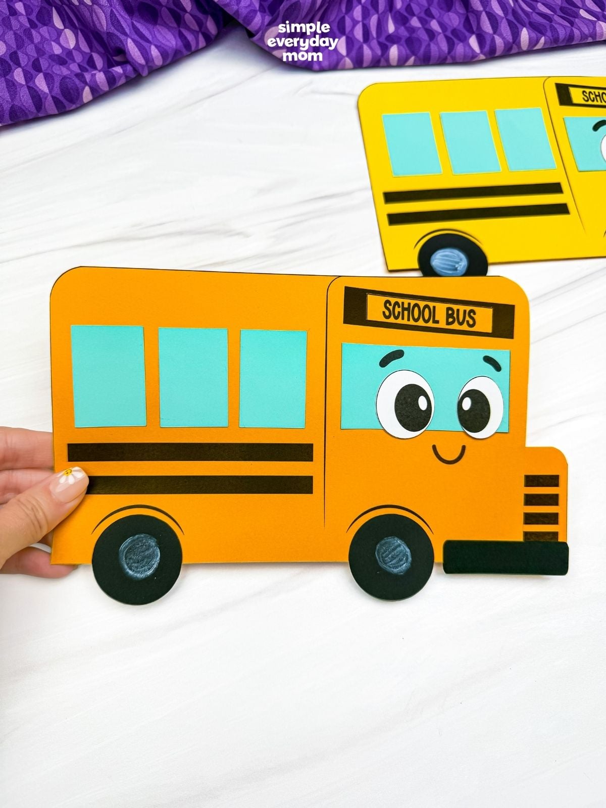 Hands holding the completed orange bus shape craft with a smiling school bus face and colorful geometric shape windows.
