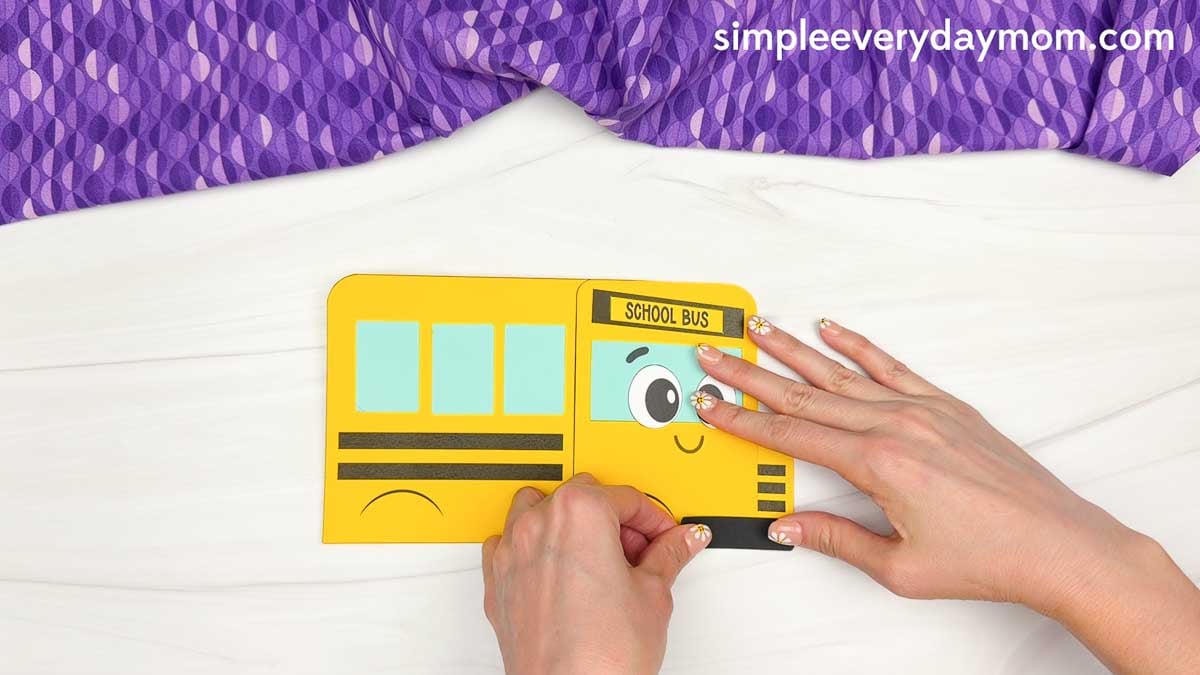 Hands attaching the bumper piece to the bottom front of the bus shape craft template.