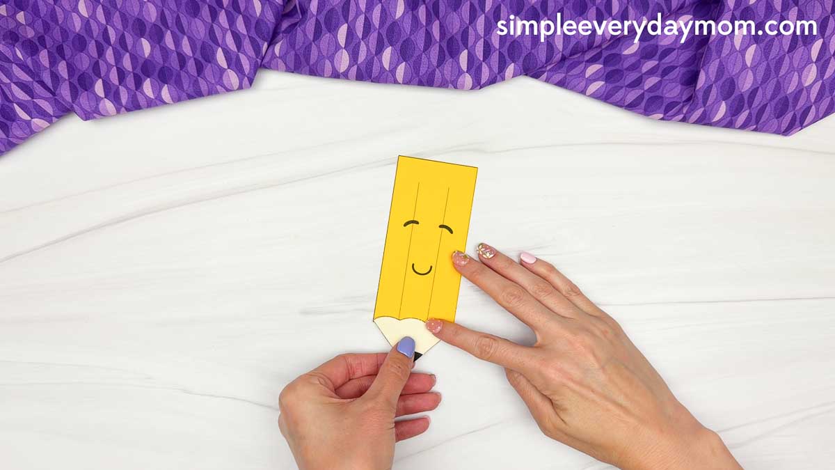 Hands attach the pencil tip piece to the bottom of the smiling pencil craft.