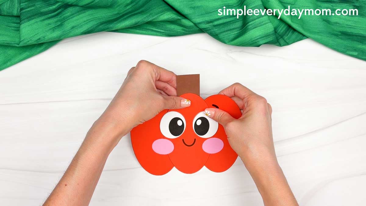 Hands gluing the stem to the top center of the Pumpkin Shape Craft.