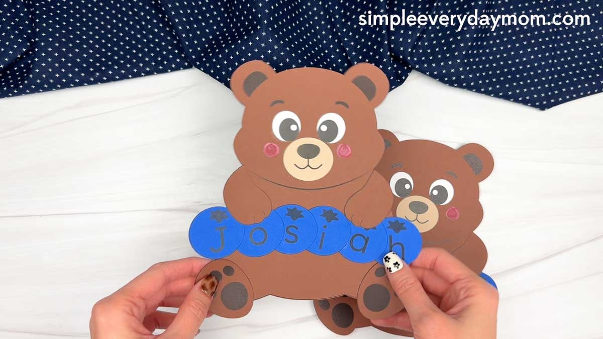 hands holding a completed brown bear name craft with blue letter pieces spelling Josiah.