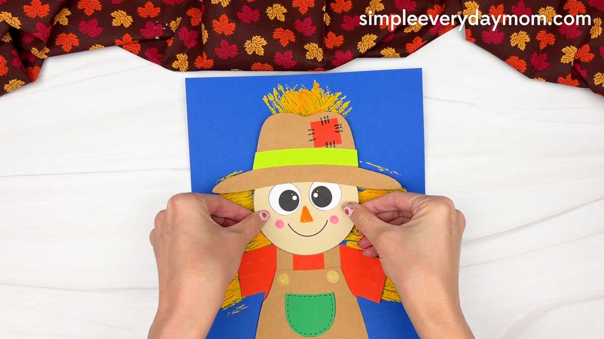 Hands gluing the scarecrow head onto the body over the painted straw background for the Fork Painted Scarecrow Craft.