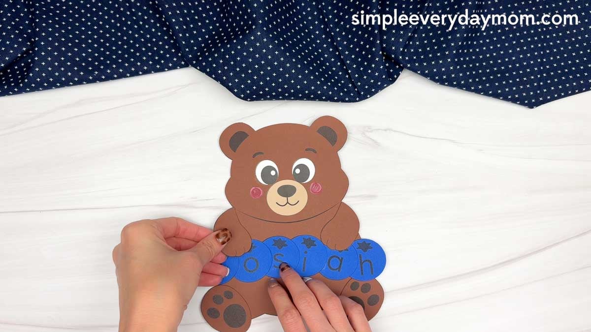 hands attaching the bear’s arms over blue letter pieces spelling Josiah.