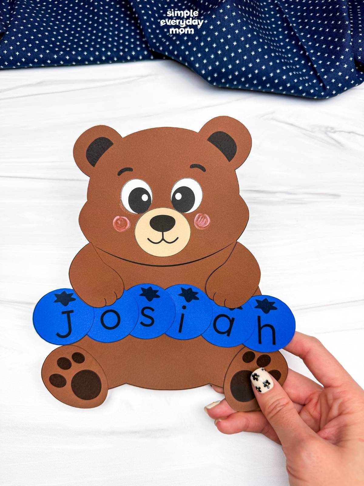 hands holding a completed brown bear name craft with blue letter pieces spelling Josiah.