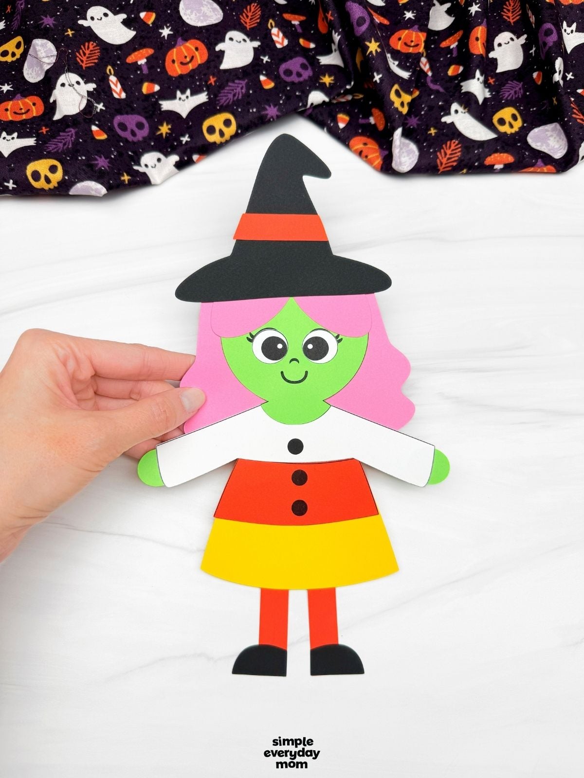 Hands holding a completed Witch Candy Corn Craft with pink hair, a witch hat, and a candy corn dress.