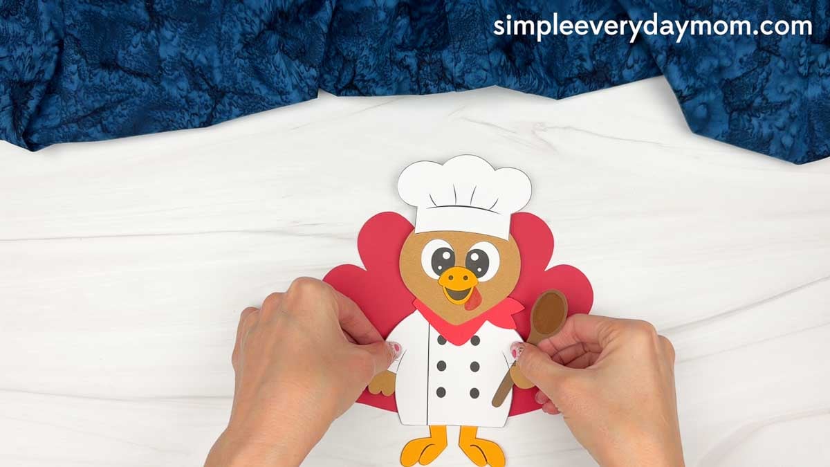 hands assembling the completed chef turkey disguise craft with red tail feathers and a brown spoon.