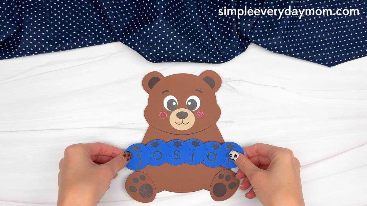 hands positioning blue letter pieces spelling Josiah across a brown paper bear name craft.