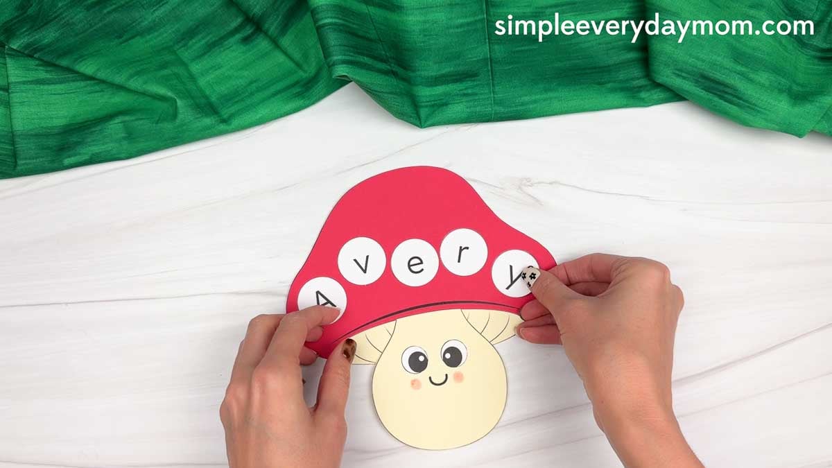 Hands adding letter circles to the red mushroom cap to spell a name for the mushroom name craft.