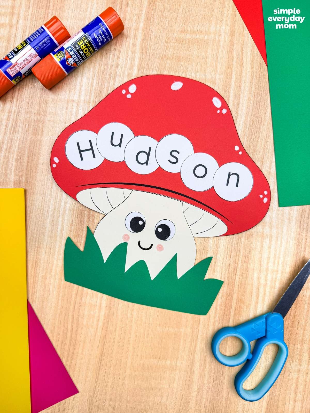 A completed Craft featuring a smiling mushroom name craft spelling Hudson, surrounded by colorful paper, scissors, and glue sticks.