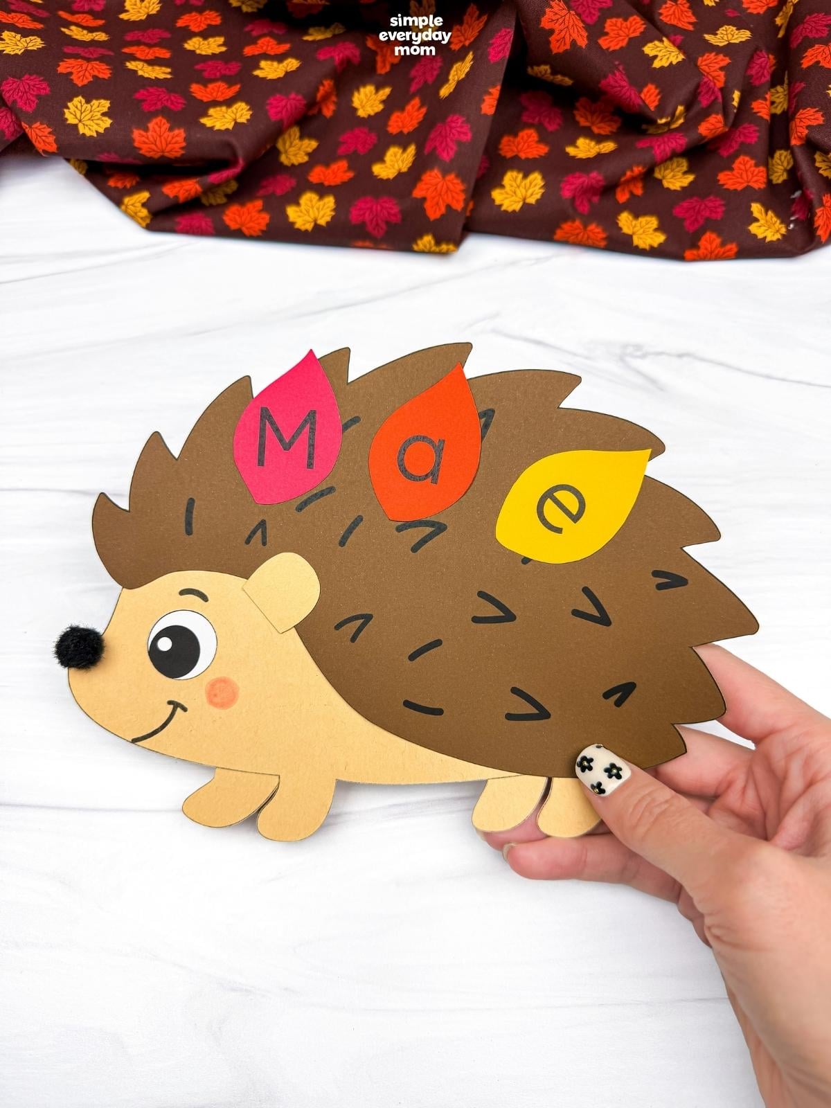 Hands holding a completed hedgehog name craft with lettered leaves spelling a child’s name.