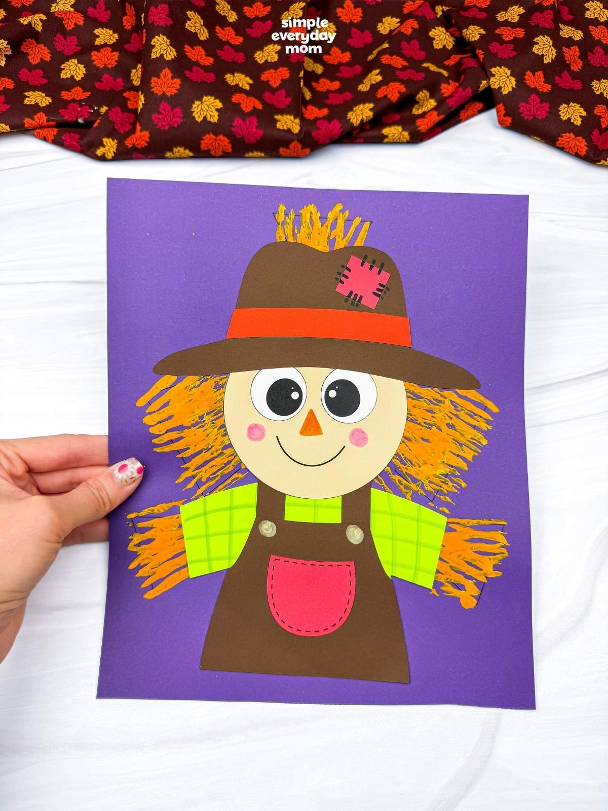 Hands holding the completed Fork Painted Scarecrow Craft with a painted straw background on a sheet of paper.
