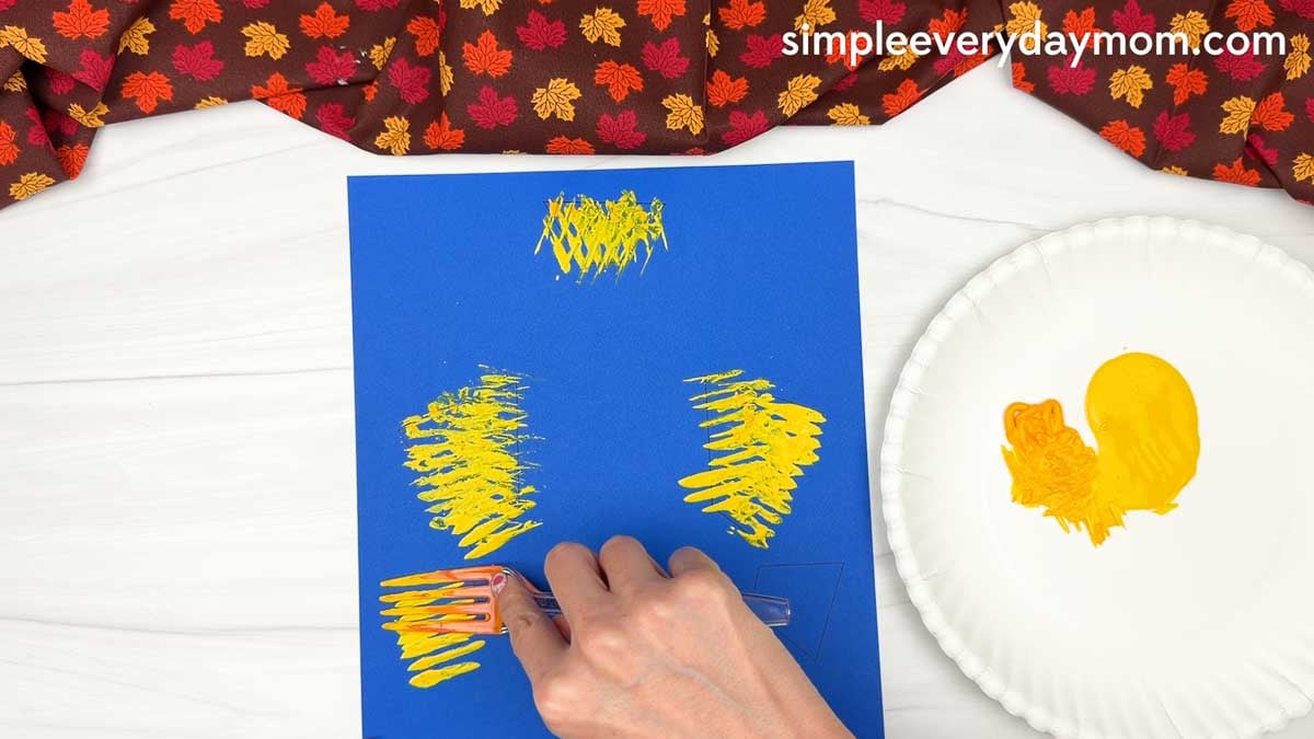 Hands using a fork to stamp painted straw onto the background paper for the Fork Painted Scarecrow Craft.