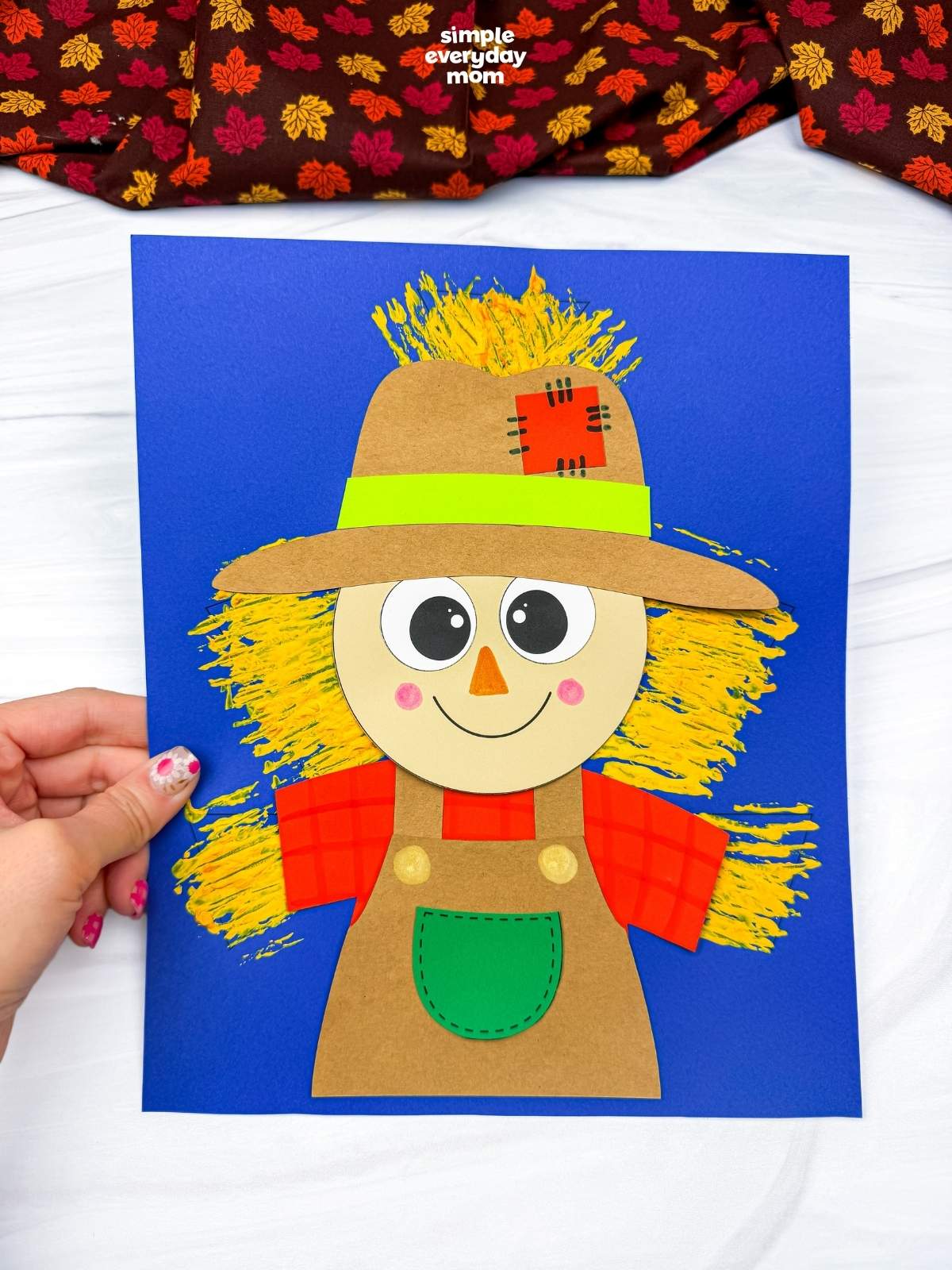 Hands holding the completed Fork Painted Scarecrow Craft with a painted straw background on a blue sheet of paper.