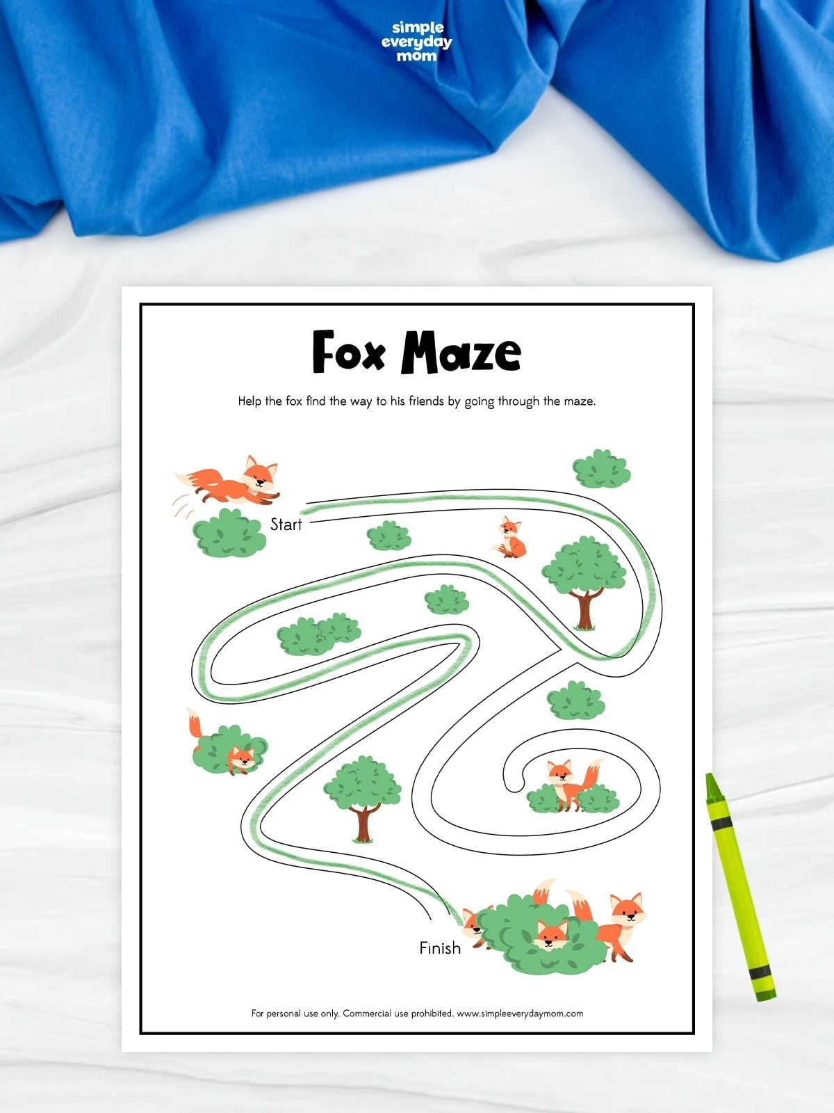 A fox maze worksheet with a winding path through bushes, trees, and fox illustrations. Children follow the maze from start to finish to help the fox reach its friends. A green crayon rests beside the completed maze activity.