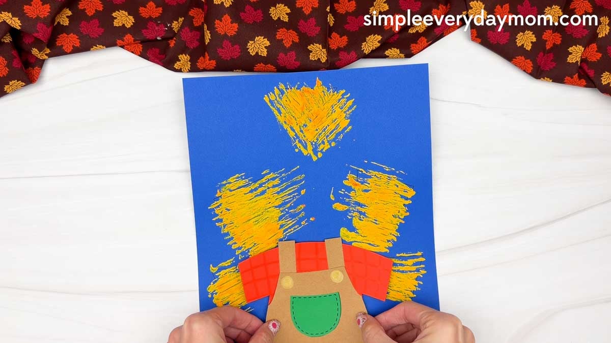 Hands positioning the scarecrow body onto the painted straw background for the Fork Painted Scarecrow Craft.