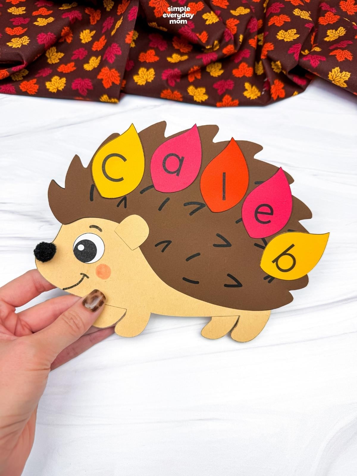 Hands holding a completed hedgehog name craft with colorful lettered leaves spelling a child’s name.